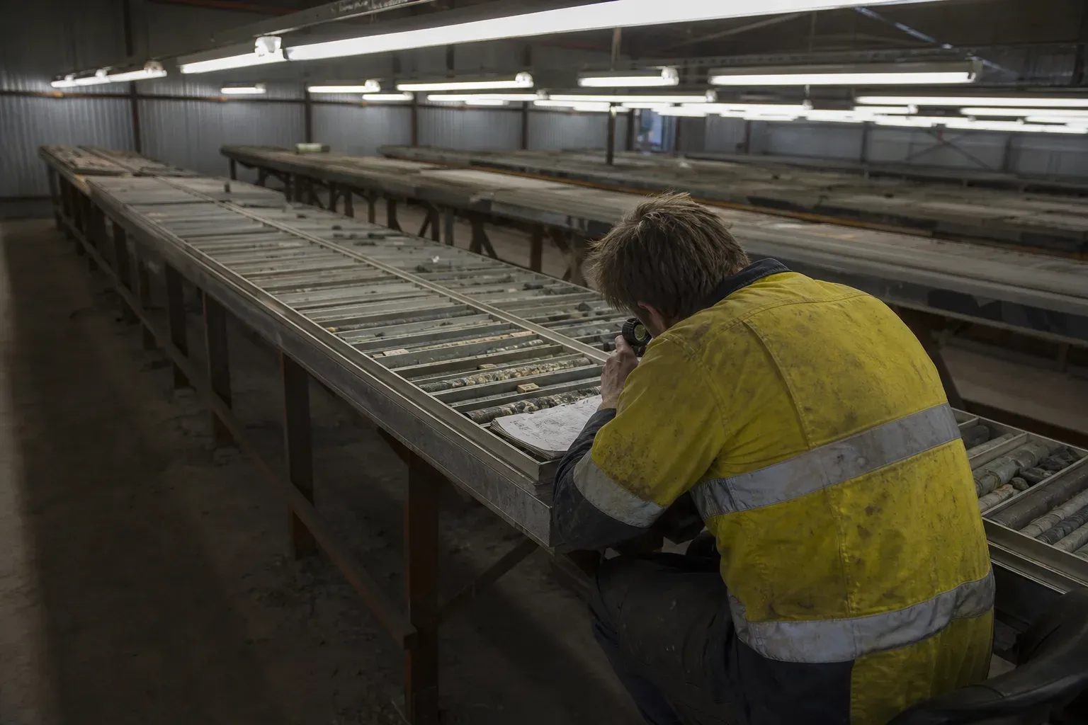 A geologist logging split drill core laid out in trays on long trestle tables in a core shed.