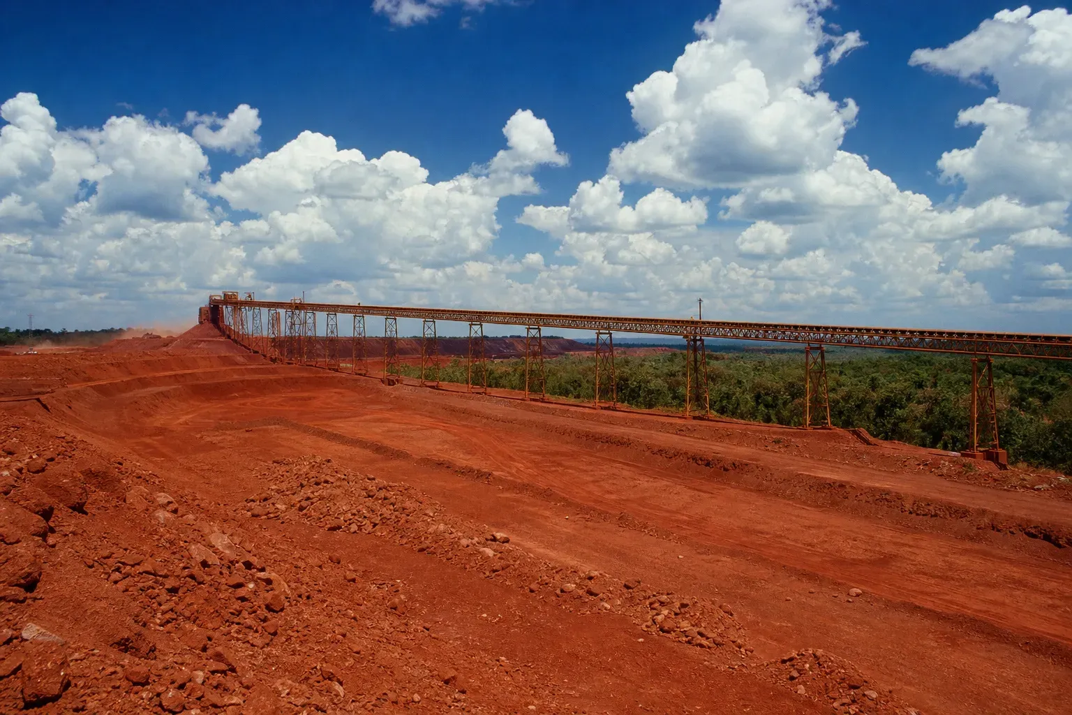 Overland conveyor on steel trestles crossing a bright red bauxite plateau under a deep blue sky with cumulus cloud.